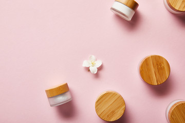 top view of scattered jars with cream and wooden caps, and jasmine flower in middle on pink