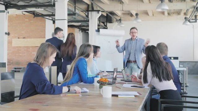 Colleagues discuss the presentation while sitting at the table. Businessman - team leader draws graphics on a flipchart and discusses business processes with employees. Office life. Coworking