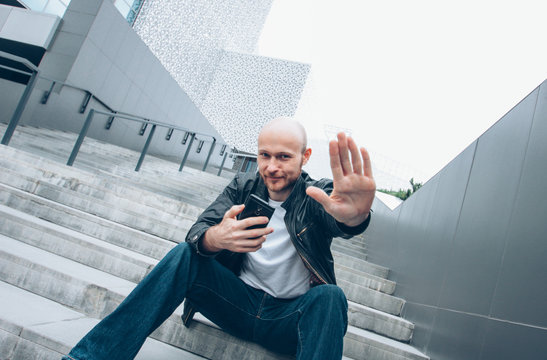 Adult bald bearded smiling man with mobile sitting in stair closing camera by hand, no more photo