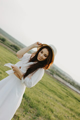 Fototapeta premium Portrait of charming carefree long hair asian girl in white clothes and straw hat enjoys life in field at sunset. Sensitivity to nature concept