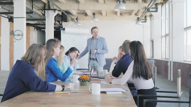 A male manager stands near a flipchart, holds a presentation and discusses with colleagues. Employees ask questions and argue. Office life. Coworking