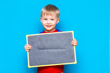small child holding chalk blackboard standing against blue background with schoolbag
