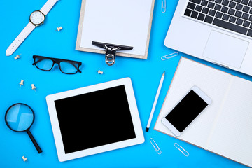 Tablet, laptop computer with glasses, smartphone and clipboard on blue background