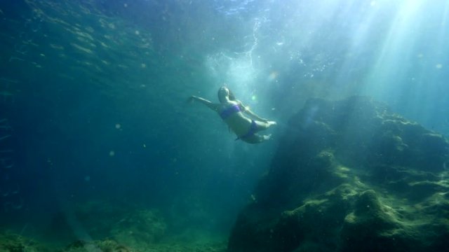 Young beautiful girl in a bathing suit floating under water on the background of a rock illuminated by the rays of the sun. She perfectly performs gymnastic figures. 