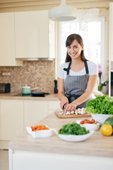 Obraz na płótnie Canvas Beautiful smiling dedicated Caucasian brunette in apron standing in kitchen and chopping mushrooms. On table are lots of vegetables. Cooking at home concept.