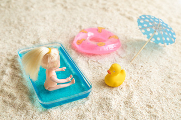 Baby girl doll sitting in kids swimming pool next to inflatable pool float, cocktail parasol and rubber duck on the beach minimal creative summer vacation concept.