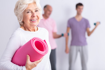 Happy senior woman holding pink joga mat, men exercising in background