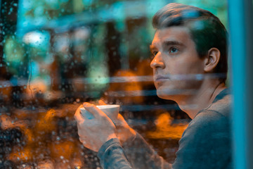Handsome man drinking tea or coffee on rainy day and looking through the window