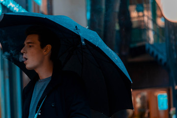 Young pensive handsome man with umbrella on rainy day walking in park.
