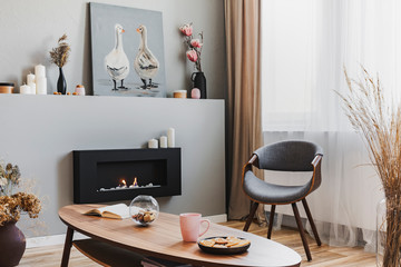 Cookies and pink team mug on wooden coffee table in grey living room interior with eco fireplace and chic chair