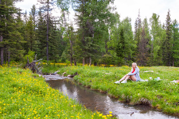 Fototapeta na wymiar A picnic on the bank of a mountain river with green grass and yellow flowers against the background of coniferous trees and a blue sky with clouds; a beautiful blonde is sitting