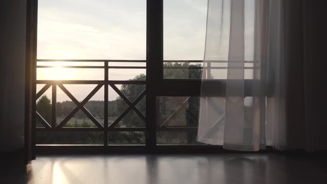Large wooden opened window with curtains waving on the wind. Beautiful summer landscape behind the balcony. Camera moving right. Sunset concept