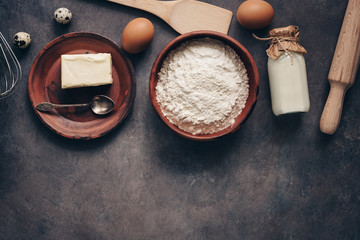 Ingredients for baking on a dark rustic background, flour, butter, eggs, rolling pin, whisk and paddle. Top view, flat lay,copy space.