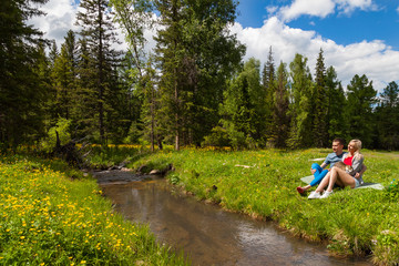 A picnic on the bank of a mountain river with green grass and yellow flowers against the background of coniferous trees and a blue sky with clouds; a beautiful blonde is sitting with man and cat