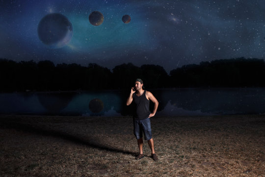 Young Man In Shorts And Cap, At Night Talking On A Mobile Phone On The Shore Of A Reservoir Against The Background Of An Abstract Night Sky.