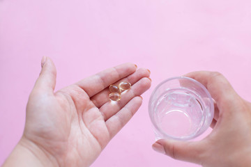 one capsule of omega three fish oil in one hand and a glass of water in the other on a pink background. taking vitamins