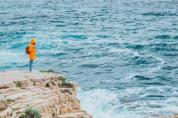 Obraz premium woman in yellow raincoat with backpack standing on the cliff looking at sea