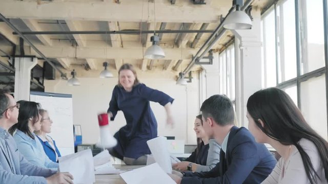 A woman manager begins to dance on the table, sings into a megaphone, throws documents and celebrates success with the team. Colleagues blow crackers confetti. Office holiday. Corporate