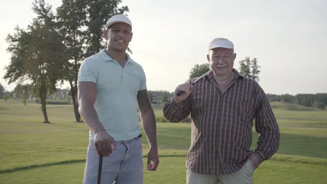 Portrait mature Caucasian man and young middle eastern man playing golf on the golf field. The golf players looking in the camera smiling. Summer leisure. Victory concept