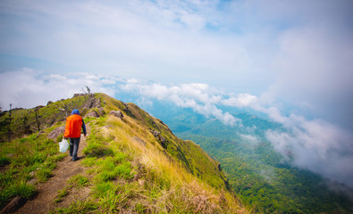 Hipster young man with orange backpack enjoying on peak mountain. Tourist traveler on blue sky and cloudy landscape background.