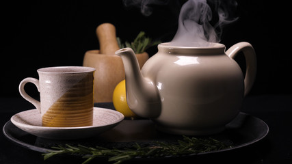 Obraz na płótnie Canvas On a black background, a brewed teapot (teapot) on a tray is a lemon and a grassy saucer, from the kettle a lot of steam comes out revealing a crush, hot water is filled and tea in the saucer in a mug