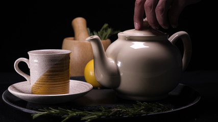 On a black background, a brewed teapot (teapot) on a tray is a lemon and a grassy saucer, from the kettle a lot of steam comes out revealing a crush, hot water is filled and tea in the saucer in a mug - Powered by Adobe