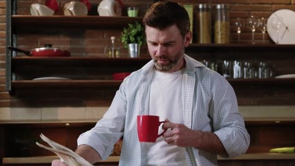 Handsome man in shirt sipping morning coffee reading news on a newspaper relaxing at breakfast in his luxury home kitchen.