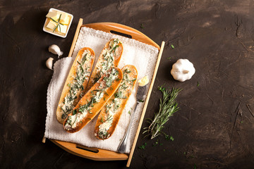 Sliced baguette toast bread with butter, garlic and herbs on a tray and dark background