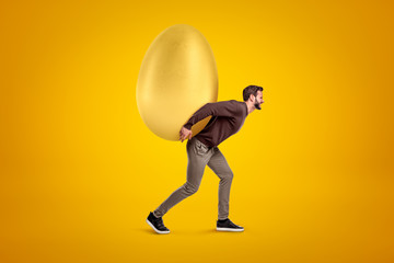 Young man in casual clothes carrying big golden egg on his back on yellow background