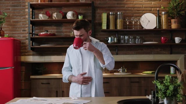 Portrait of a guy suffering from stomach pain drinking water and taking pills to stop the terrible pain standing by table in his own kitchen.