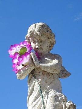 Scene in a graveyard: an old stone statue of a little angel with its hands clasped, praying. The angel holds in its arms a bouquet of artificial pink flowers. Blue sky on a sunny day.