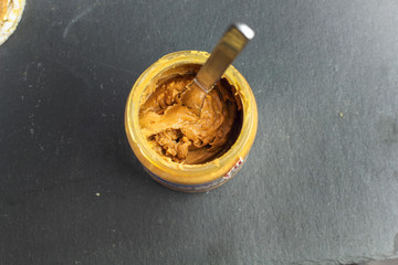 crunchy natural peanut butter in jar with knife on black slate background top view with copy space for text. Proper nutrition diet vegetarian breakfast