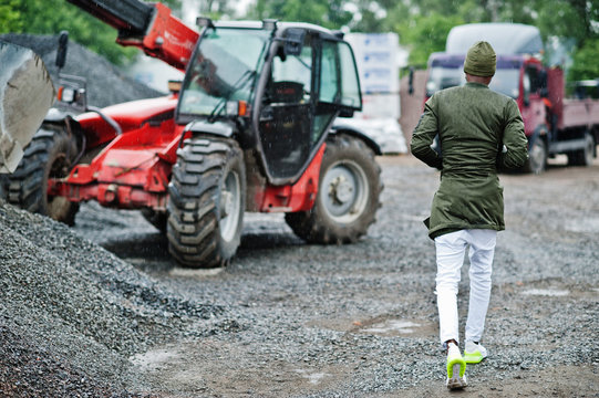 Stylish african american man in hat and sunglasses posed outdoor in rain against tractor with a bucket.
