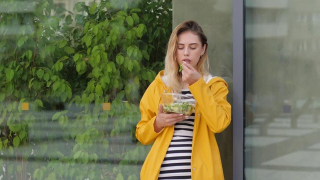 Beautiful young girl eat healthy vegetable salad from plastic bowl staying alone in yellow coat leaning a glass wall surface