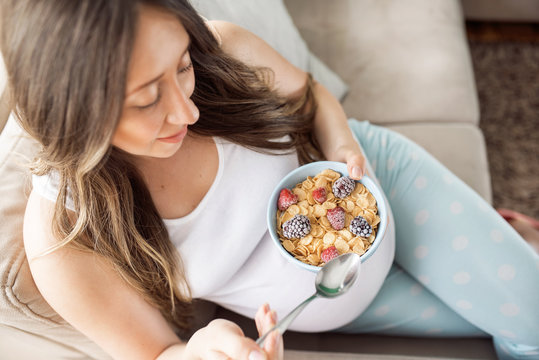 Top view of happy pregnant woman eating cereals with fruits for breakfast in bed at home