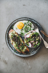 Healthy trendy breakfast. Flat-lay of avocado toast on sourdough bread with herbs and sprouts, fried egg with coriander and salad in plate over grey concrete background, top view. Clean eating concept