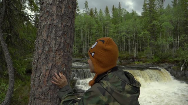 Woman touching a tree bark in a forest with a waterfall behind.