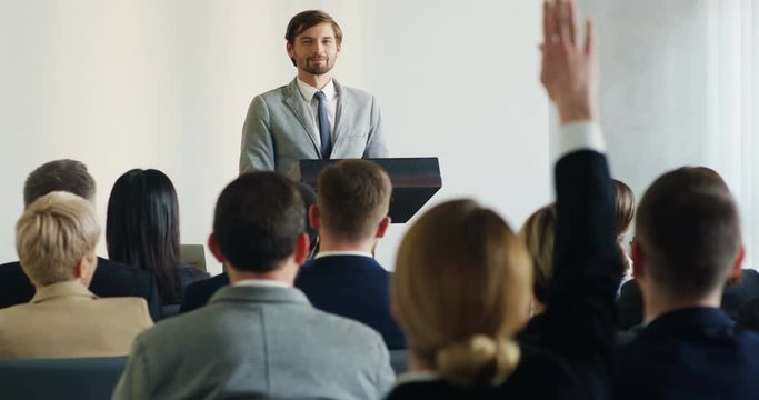 Caucasian handsome man having a speech at the business conference, pointing at the woman who raising ger hand to ask a question.