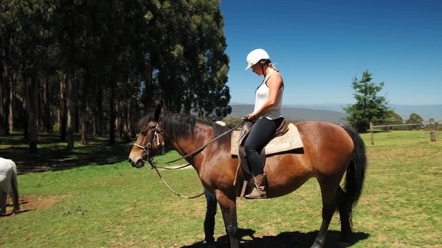 Woman sitting on horse whilst another person prepares the horse tack equipment.