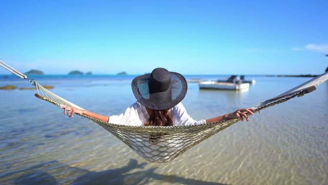 Camera behind the Elegant woman wearing black hat swings on hammock and watching beautiful seaside view