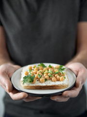 Obraz na płótnie Canvas Male hands takes plate with vegan sandwich. Healthy appetiezer - whole wheat bread toast with plant-based soft cheese, chickpea and fresh green parsley. Vertical. Copy space for text. Vertical.