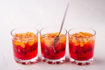 Jelly dessert with fruits apricot raspberry in three drinking glasses with single spoon yellow red summer white background isolated in row