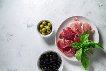 Slices of jamon and prosciutto on a white plate and two bowls with anchovies and olives on a light background. Top view with copy space for your text