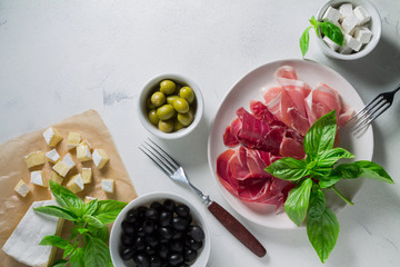 Slices of jamon and prosciutto on a white plate and bowls with anchovies, olives and feta cheese on a light background. Top view