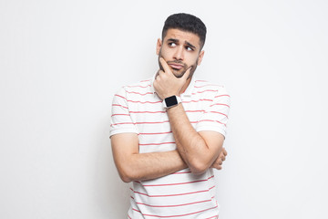 Portrait of pensive handsome bearded young man in striped t-shirt standing, touching his face,...