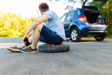 Fototapeta na wymiar Sad and depressed person sits on a spare wheel near a blue car with a punctured tire and an open trunk. A man calls using a smartphone mobile tire service.