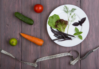 Healthy food and diet vegetarian concept - measure tape, white plate with leaf of salad, cucumber, tomato and other vegetables