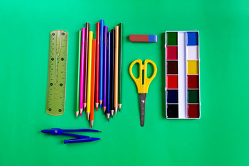 A set of paints, pencils, scissors, ruler, eraser and compasses on a green background with copy space. Collection of school supplies