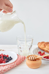 Croissants on plate on the wood board, berries, coffee, milk and honey on a white tablecloth. Male hand pours milk into a glass with coffee. Lifestyle. Copy space