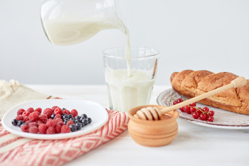 Croissants on plate on the wood board, berries, coffee, milk and honey on a white tablecloth. Male hand pours milk into a glass with coffee. Lifestyle. Copy space